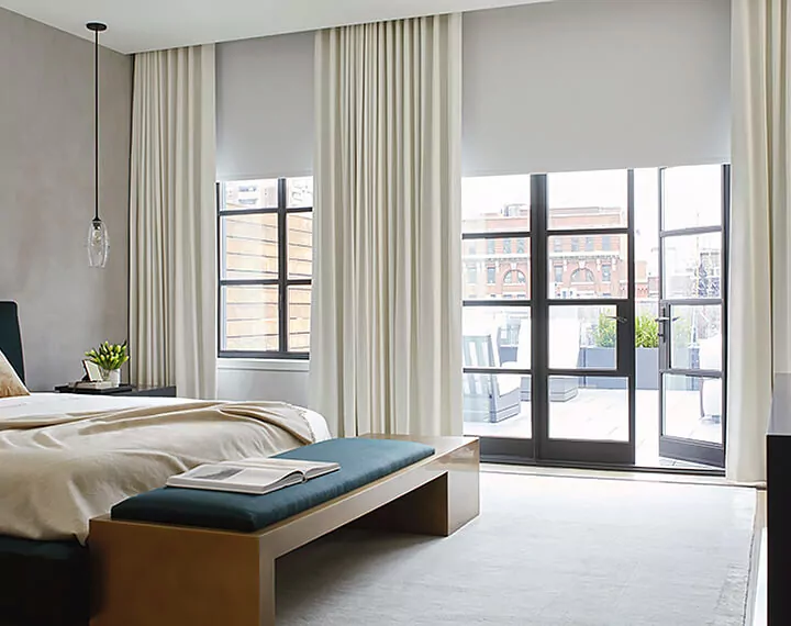 A bright bedroom with blackout Roller Shades and Drapery over large paneled windows and patio doors.