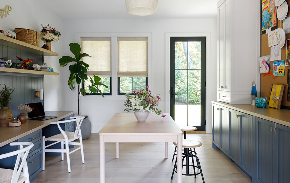 A creative workspace has earthy tones like deep blue cabinets, a wood desk and Flat Roman Shades made of Grassweave in Hemp