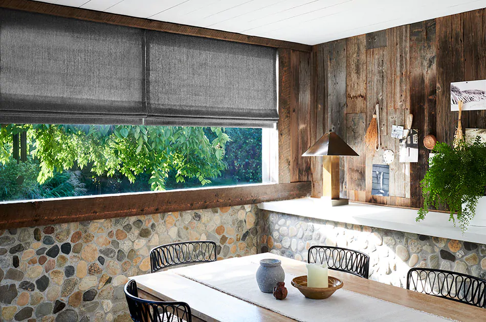 A rustic dining room with stone walls has Nate Berkus Lowell Tweed in Flint fabric for Roman Shades for a stony grey color