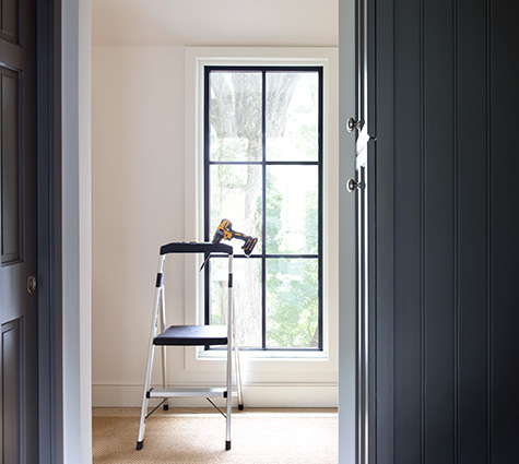 A stepladder with a drill resting in the holder sits in front of a tall narrow window with black mullions