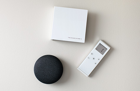 The Shade Store motorization remote and wireless link sit on a table next to a small round black Google Nest device