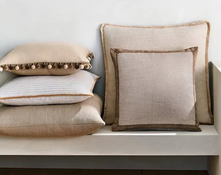 Several Square Pillows in neutral fabrics with various trim on a white bench.