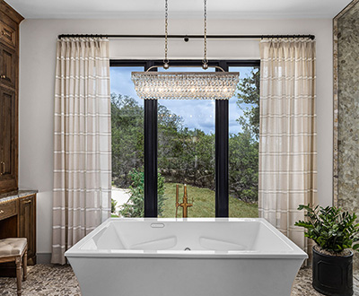 A luxe bathroom has a square marble tub and Pinch Pleat Drapery made of Victoria Hagan Harbor Stripe in Sand