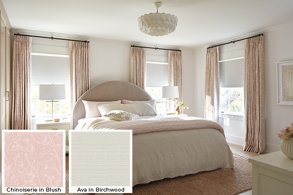A cozy bedroom with pink and white colors has blackout Roller Shades for windows made of Ava in Birchwood paired with drapery