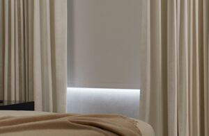 A modern bedroom has blackout Roller Shades for windows made of Cora Blackout in White pulled down for room darkening