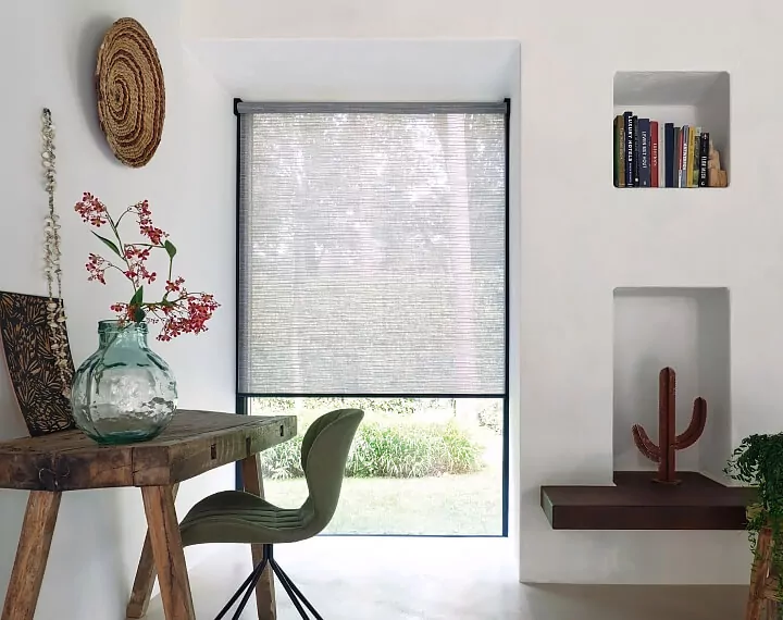 Office with a natural wood desk and a window with grey roller shades.