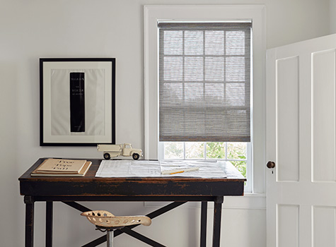 Roller Shades for windows cover a small window with a rustic wood desk in front of it with papers and pencils on top