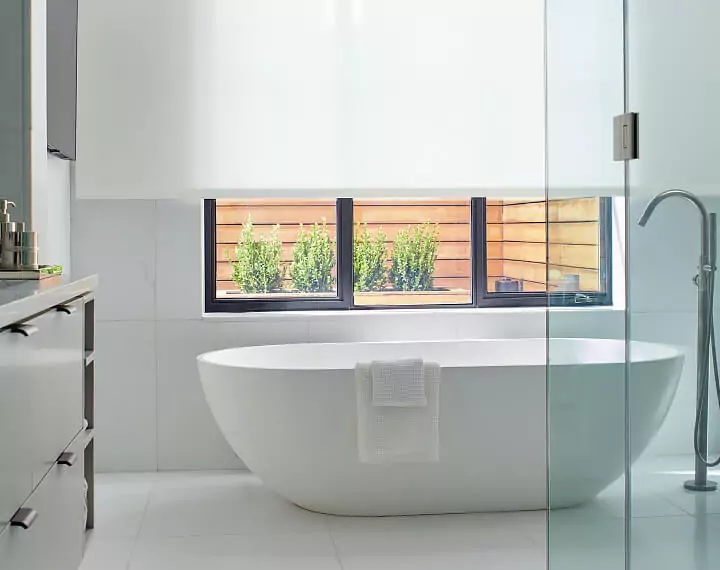 White bathroom with a bathtub and window with light filtering solar shades in white.