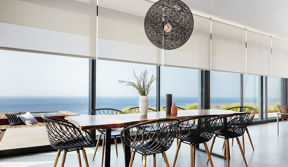 A dining table with woven chairs sits in in a sunny room with solar shades for sliding glass doors partially raised