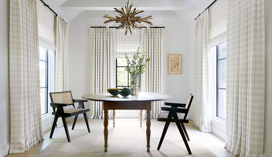 A dining room has neutral curtains layered with Flat Roman Shades all made of Holland & Sherry Emerson in Shea