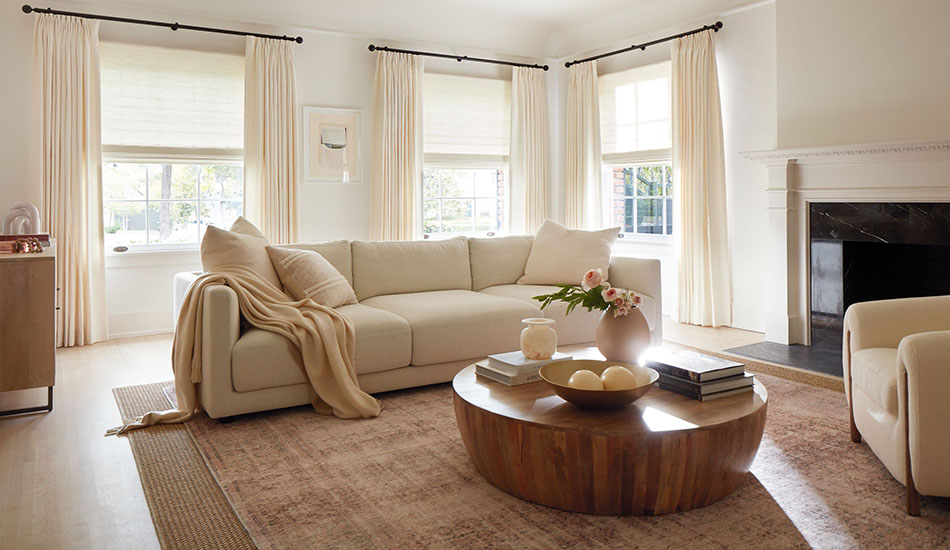 An inviting living room has neutral curtains made of Luxe Linen in Oyster paired with woven shades in Harper, Ivory