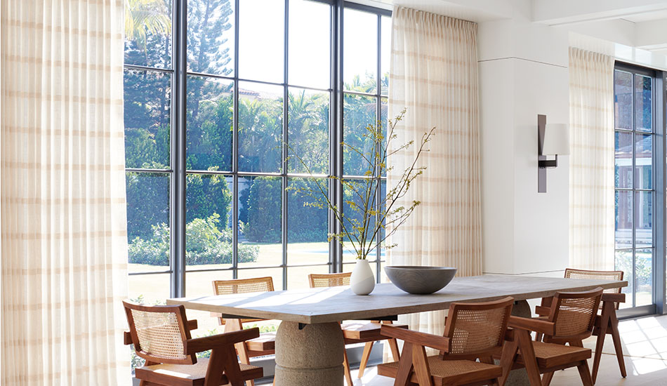 A long dining table with wood chairs sits in front of a sunny window with Tailored Pleat Drapery made of Lily in Buff