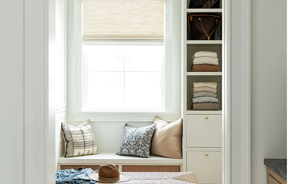 A walk-in closet has a window bench that incorporates window seat ideas like a Woven Wood Shade, throw pillows and drawers