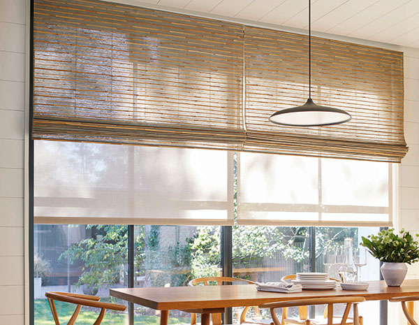 A sunny dining room has a wood table and sliding doors with layered Roller Shades and Woven Wood Shades for lots of texture